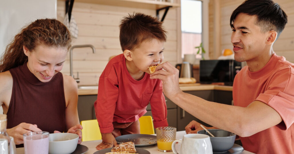Mamá y papá desayunan en la cocina mientras el papá le da de comer a su hijo pequeño.
